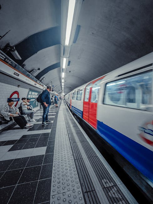 A subway station platform with a curved ceiling illuminated by long fluorescent lights. On the left, four seated passengers wait, some looking at their phones or reading. A man in a dark jacket and je