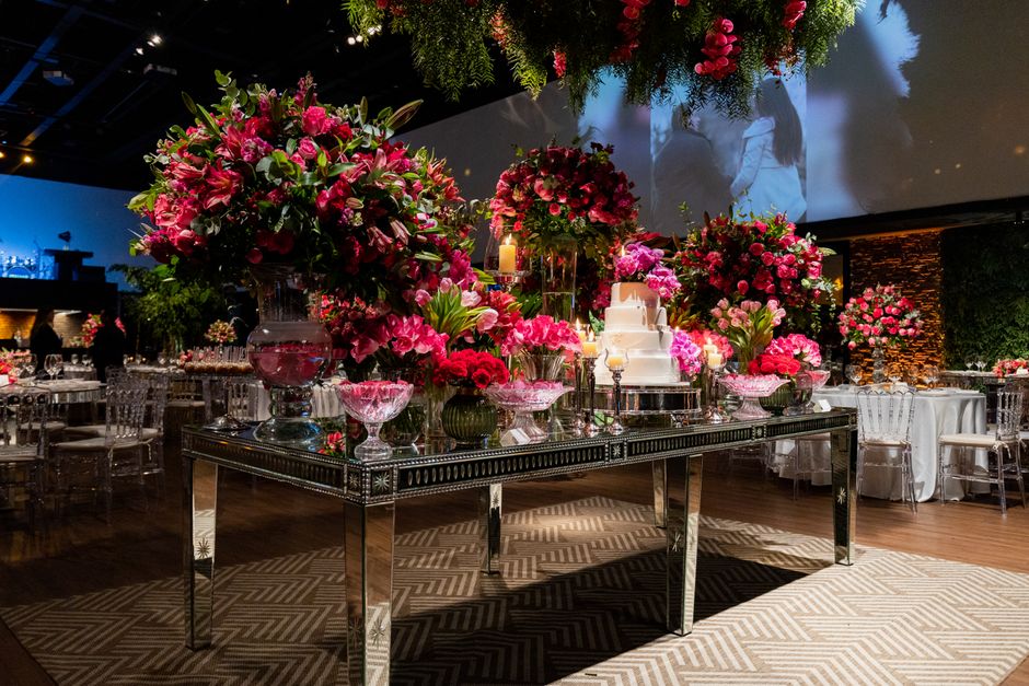 A luxurious floral display featuring large arrangements of vibrant pink roses, lilies, and peonies placed in clear glass vases on a reflective black table, surrounded by smaller bouquets of similar fl