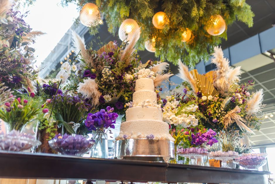 A three-tiered white wedding cake with intricate textured icing, displayed on a reflective silver stand amidst an abundant floral arrangement. The bouquet features a variety of fresh flowers including