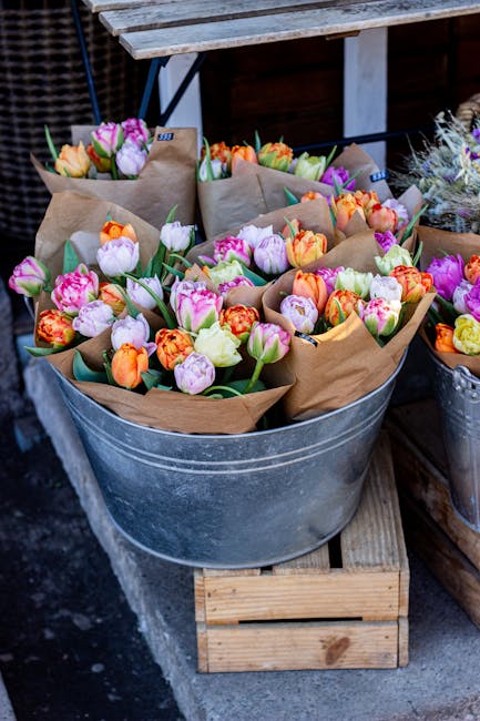 A galvanized metal tub filled with multiple bouquets of fresh tulips and ranunculus flowers, wrapped in brown craft paper and secured with green floral tape. The tulips display a variety of colors inc