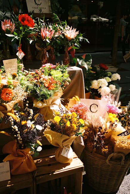 An arrangement of various fresh flowers displayed on a wooden table at a flower shop, including bouquets wrapped in brown paper and tied with ribbons. The flowers feature vibrant colours such as red, 