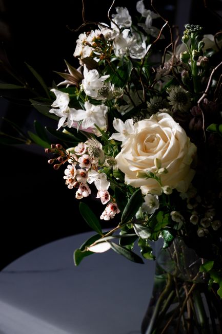 A bouquet arrangement featuring a large, creamy white rose as the centerpiece, surrounded by smaller white blossoms, pink-tinted flowers, and lush green foliage. The flowers are arranged in a natural,